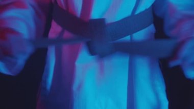 Küçük bir çocuk neon ışıkta tekvando yapıyor. Kemeri bağlıyor ve elleriyle hareketlerini gösteriyor.