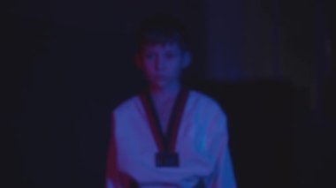 Karanlıkta tekvando yapan küçük bir çocuk neon ışıklarıyla kameraya doğru yürür ve jest yaparak ellerini gösterir.