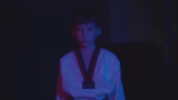 Un niño pequeño haciendo taekwondo en la oscuridad con luz de neón camina hacia la cámara y muestra sus manos en gesto — Vídeos de Stock