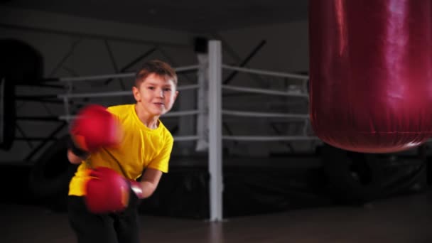 Sonriente niño en guantes de boxeador camina a un saco de boxeo y golpearlo — Vídeo de stock