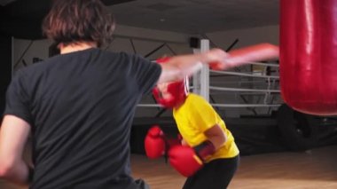 Yumuşak koruyucu kasklı küçük bir çocuk genç bir koçla boks yapıyor. Uzun yumuşak sopalara vuruyor.