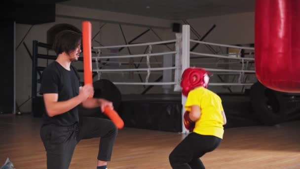 Niño pequeño en casco protector haciendo boxeo con un entrenador joven - golpeando en los palos largos y suaves — Vídeos de Stock