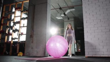 Pilates yapan genç bir kadın - fitness topuna doğru yürür - üzerine oturur ve egzersiz yapmaya başlar.