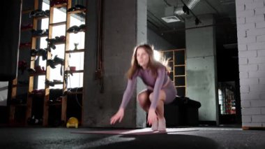 Pilates egzersizi yapan genç bir kadın yoga minderine yürür ve bölmelere oturur.