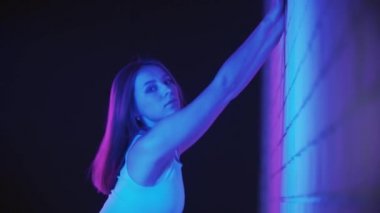 Genç bir kadın neon ışıkla egzersiz yapıyor, belini duvara yaslıyor.