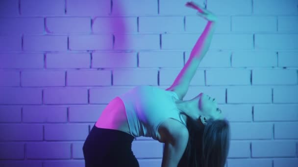 Ung kvinna tränar i neon belysning - stretcha och böja tillbaka ryggraden framför tegelväggen — Stockvideo