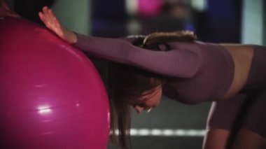Mor spor takım elbiseli genç bir kadın spor yapıyor. Ellerini spor topuna dayayıp sırtını eğiyor.