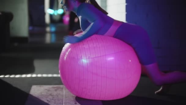 Jovem mulher de terno esportivo roxo fazendo fitness rasteja na bola de fitness e esticando os dedos dos pés até a cabeça — Vídeo de Stock