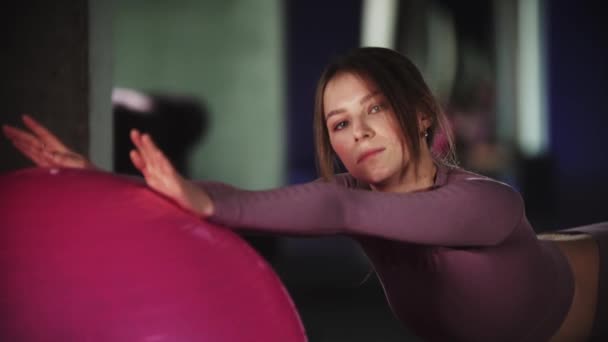 Giovane donna in tuta sportiva viola che fa fitness - appoggiandosi con le mani sulla palla fitness e piegando la schiena - intravede la fotocamera — Video Stock