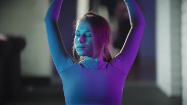Mladá hezká žena v purpurovém sportovním obleku dělá fitness - dělá cvičení na józe podložka v neonově modrém osvětlení — Stock video