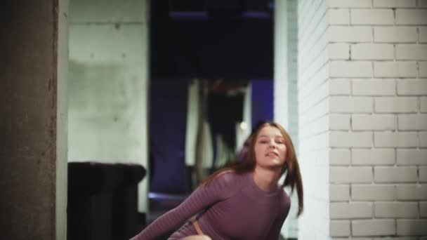 Молодая улыбающаяся женщина в фиолетовом спортивном костюме идет вперед к камере в тренажерном зале - садится на йогу и кладет волосы в хвостик — стоковое видео