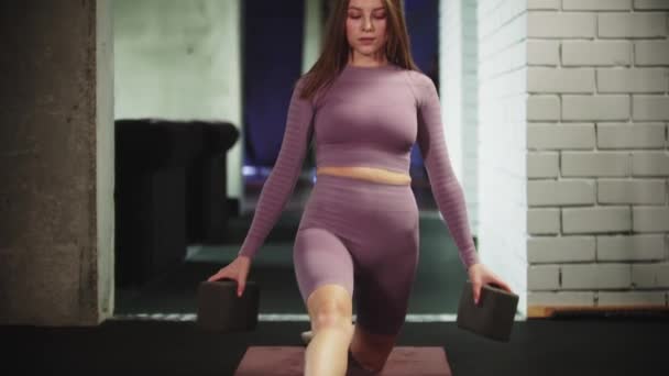 Joven mujer bonita en traje deportivo púrpura sentada en las divisiones usando soportes especiales para sus manos y se ve en la cámara — Vídeos de Stock