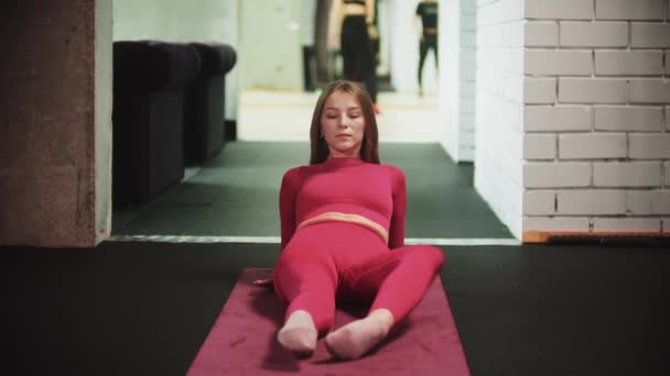 Mulher bonita nova no terno vermelho dos esportes que faz esportes no gym - deitar-se no tapete do ioga e exercitar suas pernas — Vídeo de Stock