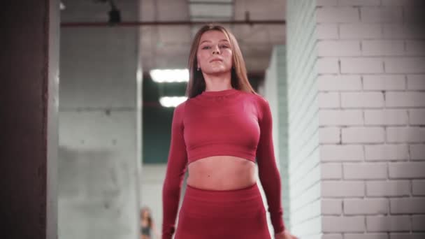 ジムでフィットネス演習を行う赤のスポーツスーツの若い女性-カメラを見て、彼女の体と前方に曲げ — ストック動画