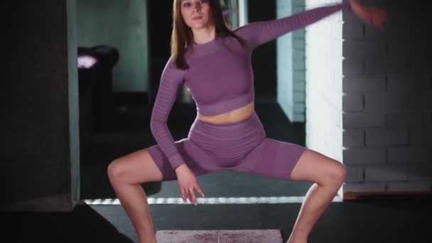 Jovem mulher em terno esportivo roxo fazendo agachamentos e inclinando-se para os lados com seu corpo — Vídeo de Stock