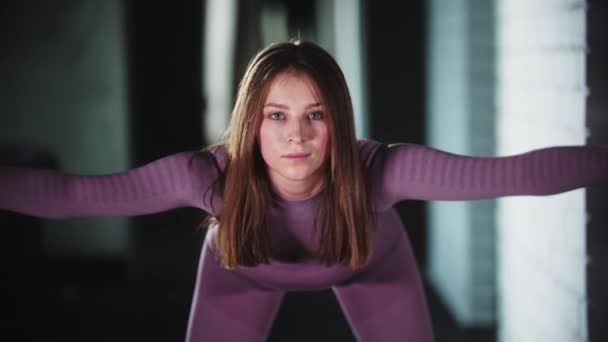 Young woman in purple sports suit doing fitness - stands feet apart and leans forward - looks in the camera — Stockvideo