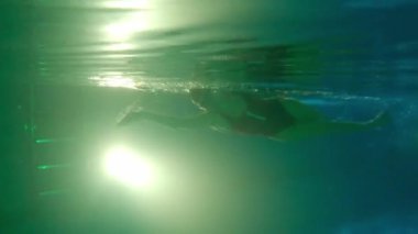 Kırmızı mayo giymiş genç bir kadın suyun altında yüzüyor. Ayaklarını yana doğru itiyor ve piste geri dönüyor.