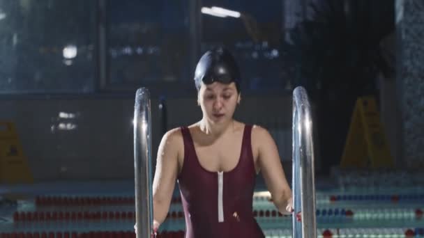 赤い水着の若い女性の水泳は、はしごを使用してプール内の水から出てくる — ストック動画