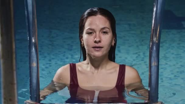 身穿红色泳衣的年轻女子站在游泳池的梯子旁边，跳入水底 — 图库视频影像