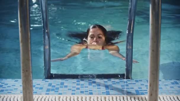 Спорт - молода жінка плаває в червоному купальнику піднімається з води в басейні за допомогою драбини — стокове відео