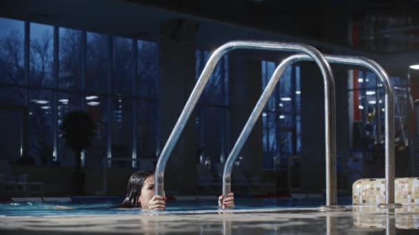 Jonge vrouw zwemmer in rood zwempak komt uit het water in het zwembad met behulp van de ladder — Stockvideo