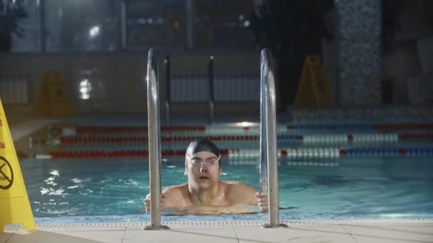 Esportes de natação - o jovem nadador sai da piscina usando a escada - ergue os óculos e olha para a câmera — Vídeo de Stock