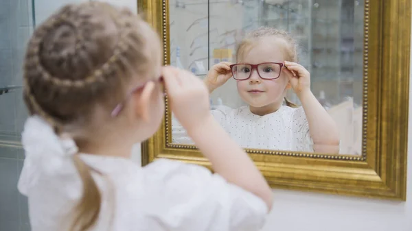 Little girl tries new glasses near mirror - shopping in ophthalmology clinic