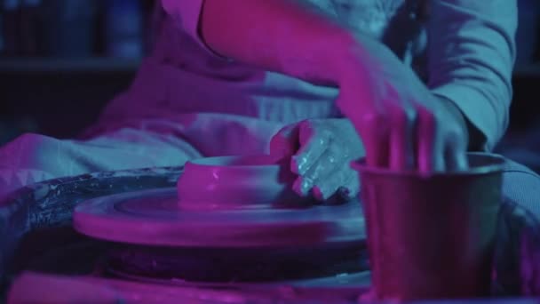 Stüdyoda neon ışıkları altında çalışan genç bir çömlekçi ellerini ıslatıyor. — Stok video