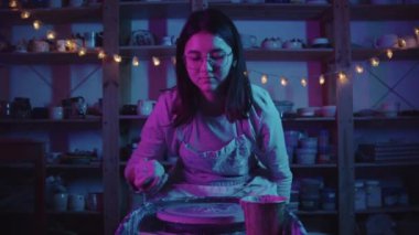 Stüdyoda neon ışıkla çalışan genç bayan çömlekçi, çamuru direksiyona koyar ve çalışmaya başlar.