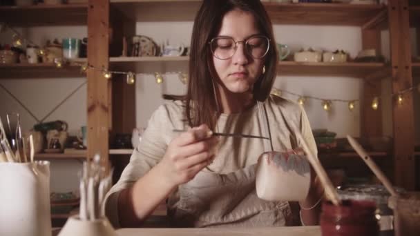 Young woman potter drawing a design on the ceramic mug with a brush — Stockvideo