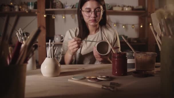 Mujer joven alfarero dibujo de un diseño en la taza de cerámica con un cepillo y luego se ve en la cámara — Vídeo de stock