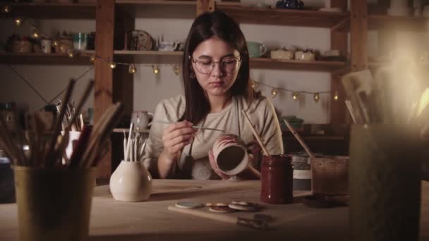 Mujer joven alfarera en vasos dibujando un diseño en la taza de cerámica con un cepillo — Vídeo de stock