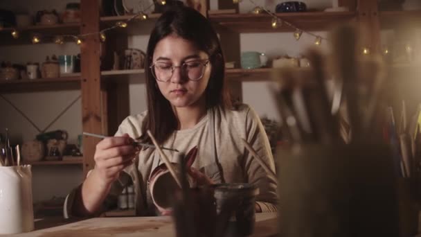 Gözlüklü genç kadın sanat stüdyosunda fırçayla seramik bardağa bir tasarım çiziyor. — Stok video