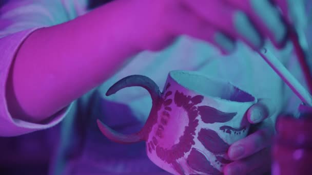 若い女性の陶芸家は、アートスタジオでブラシでセラミックマグカップのデザインを描く-ネオン照明 — ストック動画