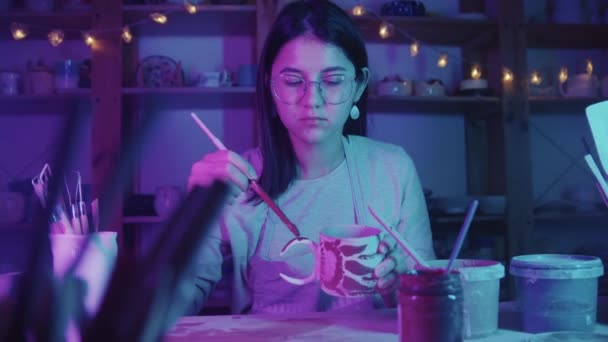 Mujer joven alfarera en gafas de pintura en la taza de cerámica con un cepillo en el estudio de arte - iluminación de neón — Vídeos de Stock