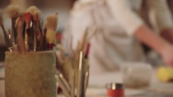 Ung kvinna keramik arbetar i konststudio - knådar lera på bordet - borstar och andra verktyg på förgrunden — Stockvideo