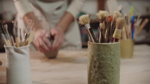 Female potter kneads clay on the table in art studio - brushes and other tools on the foreground — Stock Video