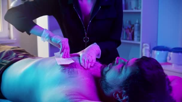 Procedura woskowania - mistrz woskowania klatki piersiowej swojego męskiego klienta paskami - oświetlenie neonowe — Wideo stockowe