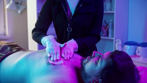 Vaxning förfarande - den kvinnliga mästaren vaxa bröstet på sin manliga klient med remsor - neon belysning — Stockvideo