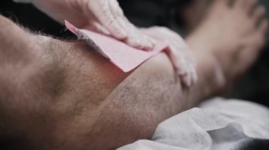Kovma ustası, erkek müşterisinin bacağındaki kılları balmumu şeritleri kullanarak alıyor.