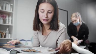 Sarışın tüy dökücü ustası, kadın müşterisinin bacağındaki kılları alırken kanepede uzanıp dergi okuyor.