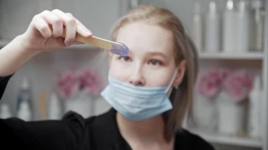 Sarışın tüy dökücü ustası spatuladaki sıcak mor balmumuna bakar sonra da kameraya bakar.