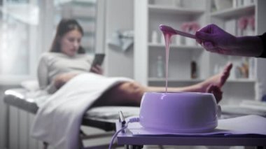 Depilasyon ustası ısıtıcıdaki işlem için balmumu eritiyor ve spatula ile karıştırıyor. Arka planda bir kanepede uzanmış telefona bakan genç bir kadın.