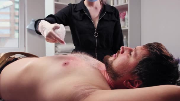 Процедура воску - бородатий чоловік лежить на дивані і майстер очищає шкіру на грудях від залишків воску — стокове відео