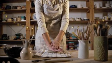 Young woman potter puts the clay on the table and using a rolling pin to flatten it