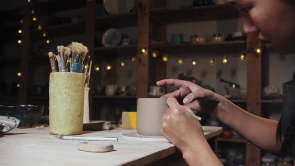 Young woman potter drawing a design on finished clay cup using an instrument — Stock Video