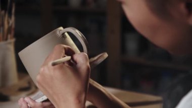 Young woman potter putting on the design to a piece of clay on the edge of the handle and the cup
