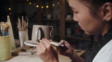 Young woman potter smears the clay on the foundation on the cup handle using a tool
