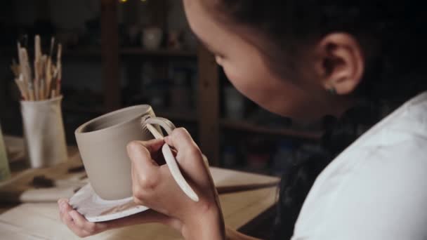 Young woman potter smears the clay from the cup to the foundation on the handle — Wideo stockowe