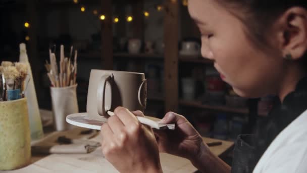 Young woman potter smears the clay on the foundation on the cup handle using a tool — Stok video